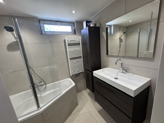 Badkamer met bad en douchescherm, handdoekradiator en donker badmeubel